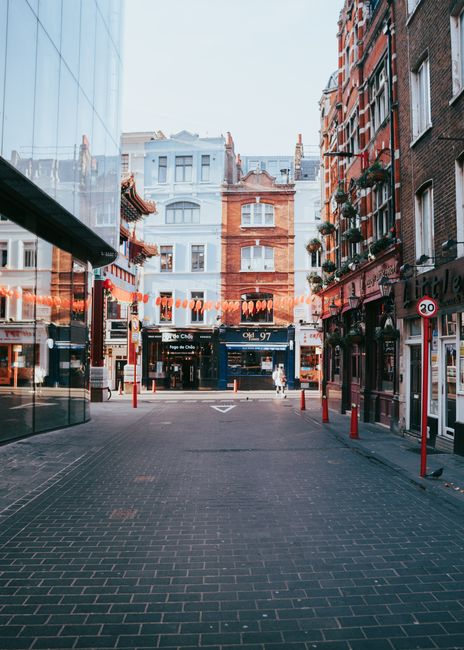 Nighttime view of a narrow cobbled street in Soho, illuminated by strings of warm outdoor string lights hanging above. The street is lined with multi-story brick buildings featuring white-framed windo