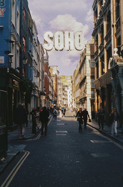 The image depicts a narrow urban street scene in Soho, Old Compton Street, with a mix of historic and modern multi-storey buildings on either side. The buildings are constructed from brick and stone,