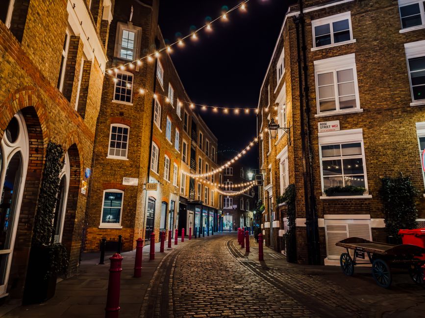 A view of a narrow urban street in Soho W1, showing a cobbled pedestrian area bordered by multi-storey buildings with brick and glass facades. On the right side, there are storefronts with signage and